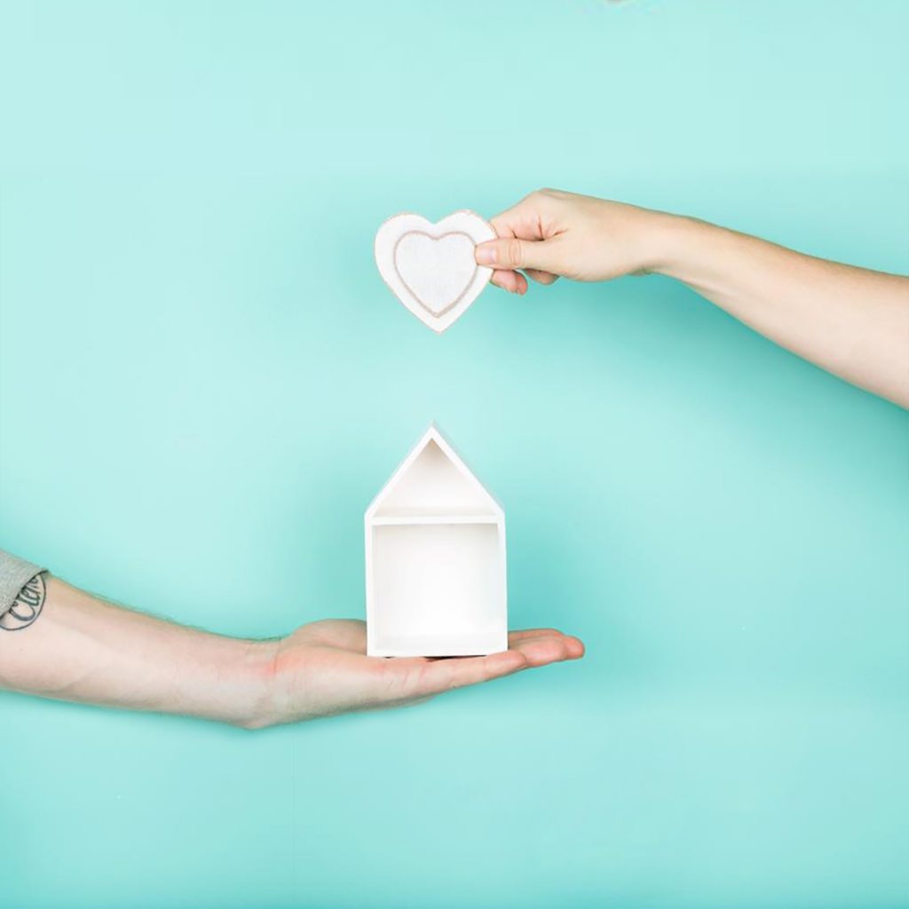 Two hands holding a heart shaped house on a turquoise background.