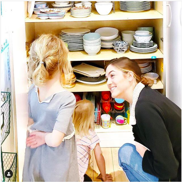 A woman supervises her children as they examine a cupboard full of dishes.