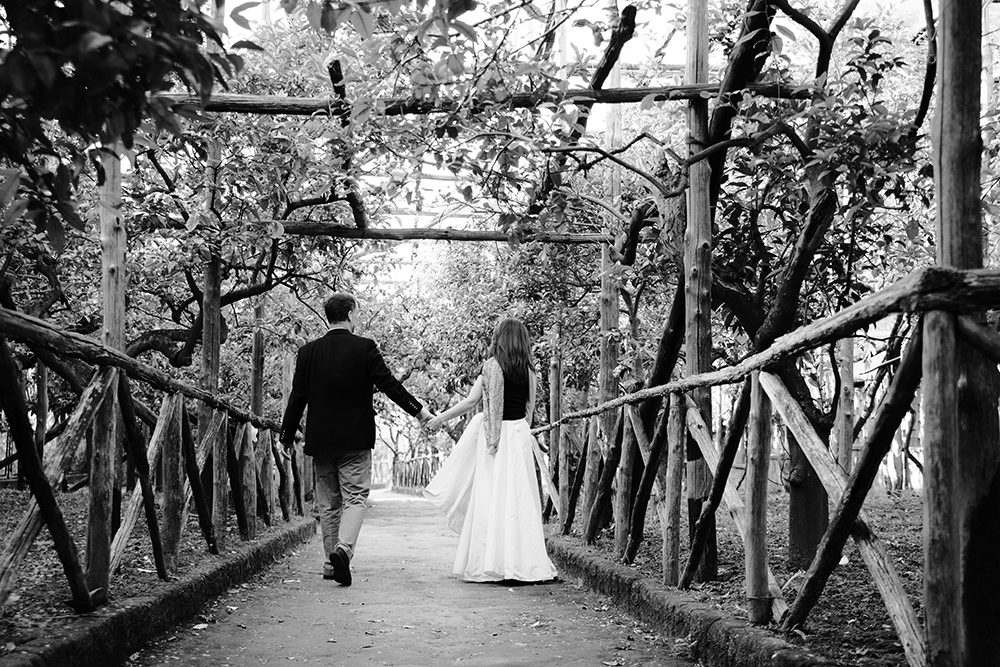 Black and white wedding photo of a bride and groom walking through an arboretum.