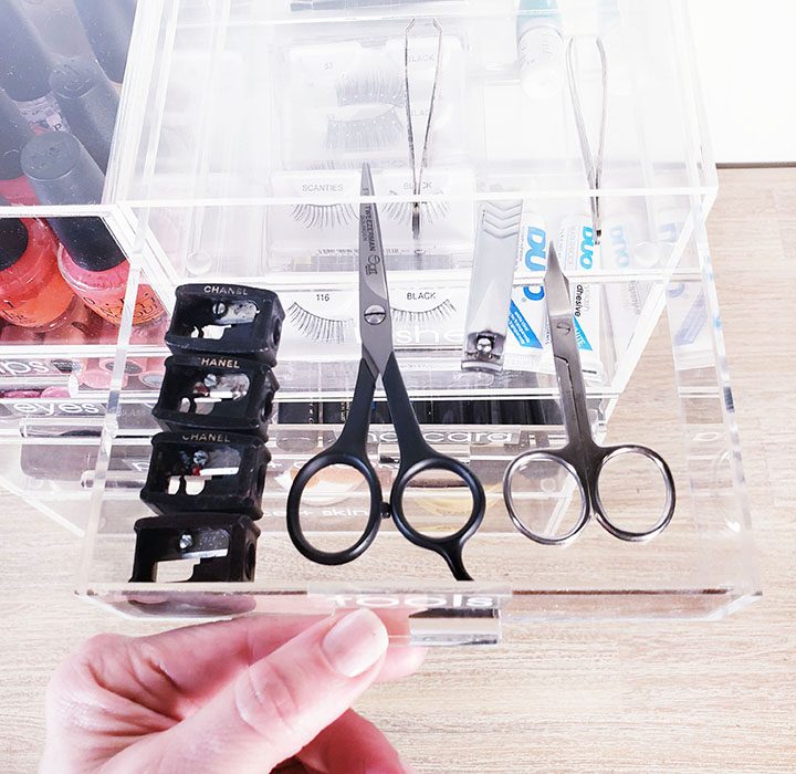 A hand holding a clear plastic box with scissors and other items in it, perfect for organizing your makeup.