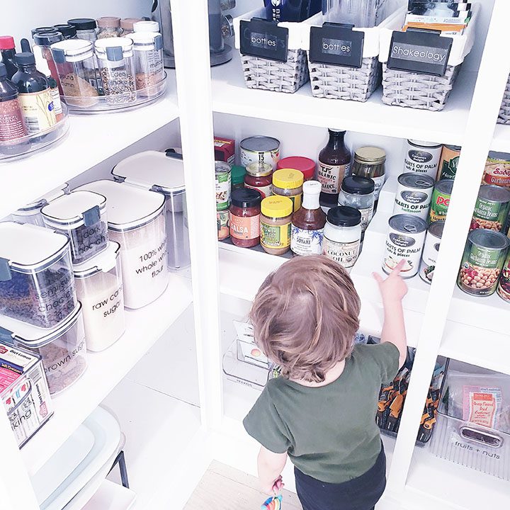 A child is standing in front of a pantry full of food.